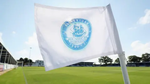 In the foreground is a white corner flag, with the blue Billericay Town FC crest printed on it, which includes a drawing of the Mayflower ship. Behind is the turf of a football pitch, with terraces. There is blue sky and white clouds above.