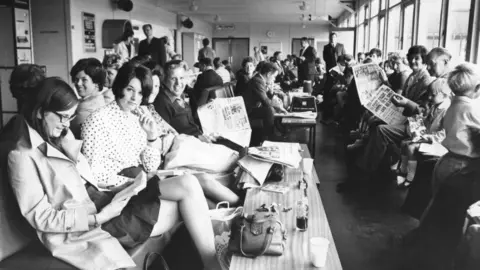 Getty Images A black-and-white image shows people sitting on rows of chairs reading newspapers, with their bags on tables in front of them. There are men, women and chidlren pictured. Some are smiling while some look fed up.