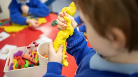 A child plays with yellow Play-Doh in their hands, sitting over a red table, with another child in the background out of focus. The photo is taken from behind the child's shoulder.