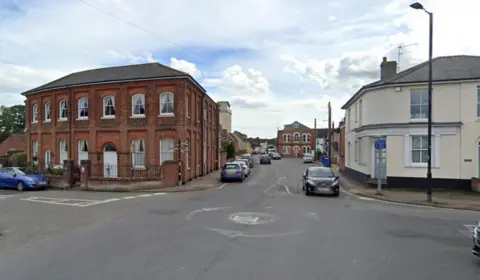 A street view of a small mini-roundabout intersection in a UK town, featuring a prominent two-story red brick historical building on the left corner and cars parked along the road stretching into the background under a partly cloudy sky.