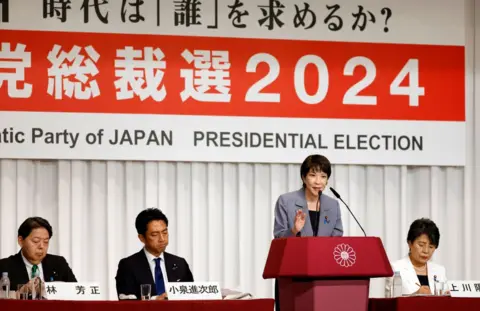 Getty Images A woman in a suit speaks at a lectern in front of a sign saying '2024 presidential election' with Japanese characters. Three other people in suits, two men and a woman, sit alongside her.