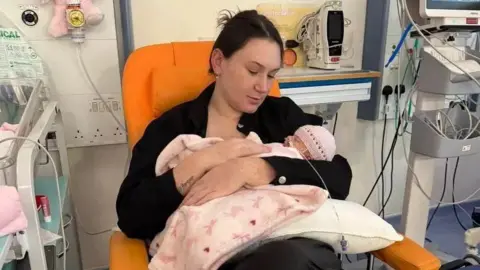 Caitlyn Gibson A mother who is brunette and in a black jumper, sat in an orange hospital chair and cradling her baby in her arms. The baby has a pink crotchet hat on and a pink blanket over her. The baby is poorly, with tubes in its nose. 
