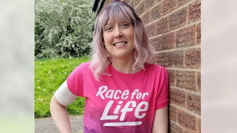 Handout Vicky Green stands against the wall of a house, with a garden behind. She is smiling and has pink shoulder length hair and is wearing a pink Race For Life T-shirt.