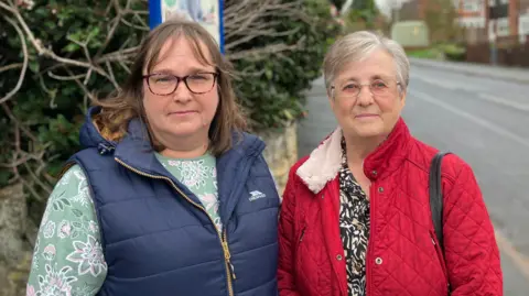 Shariqua Ahmed/BBC Julia Cunnington, a woman wearing a green floral top and blue sleeveless jacket and Jean Jackson, a woman wearing a red coat with grey short hair, stand together at a bus stop.
