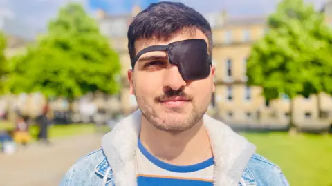 BBC Jabar Barzanji - a man with dark hair has a patch over his left eye. He wears a denim jacket and stands in front of a small park