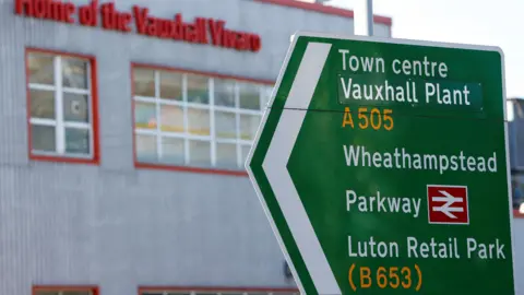 A green road sign points to the left. It says Town centre Vauxhall Plant A505, Wheathampstead Parkway and Luton Retail Park (B653). In the background is building which says in red writing Home of the Vauxhall Vivaro.