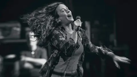 Lytham Festival Alanis Morissette in a black and white photo, singing on stage with her eyes closed and her long hair streaming behind her