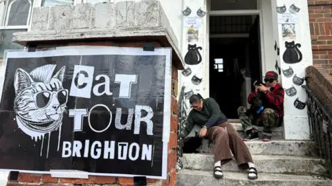 Danielle Malgwi / BBC Two cat owners and volunteers for Brighton Cat Tour sit on concrete steps outside their flat. On the red brick column in front of the steps and doorway sits a punk-themed poster reading "Cat Tour Brighton", depicting a cat with a mohawk, sunglasses and chains sticking its tongue out. The owners sit and pet two black cats, with black cat-themed decorations adorning the front entryway to their home for passing visitors.