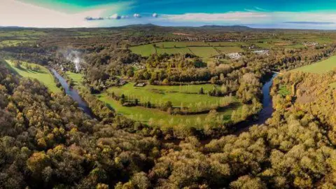 Shropshire Council A wide aerial view of a large park. The river Severn is curving through the middle of it, with thousands of trees in clusters on either side, as well as green fields. In the distance there are tall hills and white clouds.
