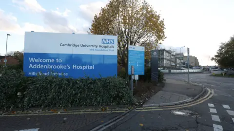 Reuters The outside of Addenbrooke's Hospital. In the foreground is a sign welcoming people to the hospital's Hills Road entrance. In the background are hospital buildings.