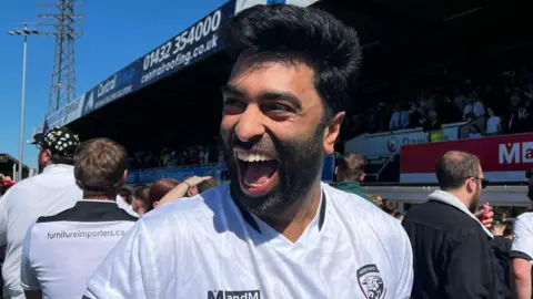 A man wearing a white Hereford FC jersey celebrates on the pitch at Edgar Street with other football fans around him.