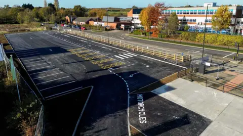 NELC A rectangular black tarmac car park with white-marked parking bays. Opposite is a school and there is a road in between.