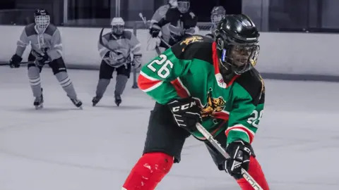 A Kenya Ice Lions player, wearing a green and black shirt, black helmet and black mittens, is pictured holding an ice hockey stick with fellow players greyed out in the background.