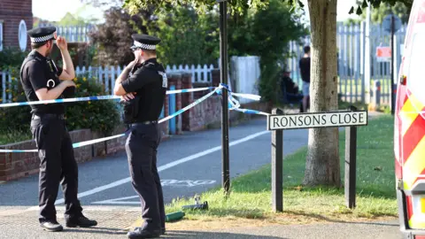 Island Echo Two policemen near a police tape at Ronsons Close. It is sunny.