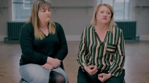 Two women are sitting on chairs and talking into a camera. One on the left is wearing a dark green top and a blue denim skirt with a slit down the middle. She has shoulder-length blonde hair and a fringe. The woman on the right is wearing a green, cream and black striped blouse and has shoulder-length blonde hair with a parting.
