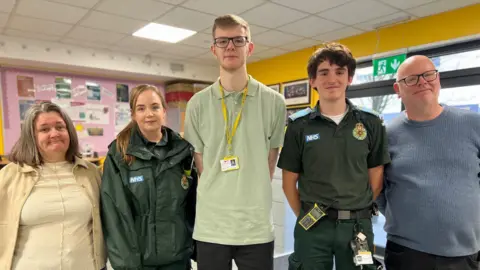 Five people stand side by side in a brightly lit classroom. Two are wearing green NHS ambulance uniforms, one person in the centre wears a pale green polo shirt with a lanyard, and two others are dressed casually. They face the camera and smile.
