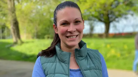 Stuart Woodward/BBC Borbala Anderson, wearing a blue t-shirt and a green bodywarmer jacket on top. She has dark brown hair scraped back into a pony tail, and is smiling for the camera. She is stood in front of a backdrop of trees and grass