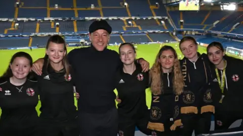 Liam Sully Media Six members of the women's team, wearing their matching kits, stand next to John Terry, who wears a black cap and jumper. They all smile at the camera with their arms around each other, at Stamford Bridge football ground. Blue seats and the football pitch are seen behind them.