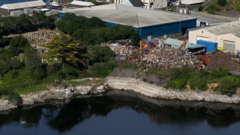 A drone shot showing the proximity between the Guernsey Scrap Metal, the St Sampson's Parish church and Longue Hougue reservoir. Scrap is falling from a large mountain of waste into the quarry. 