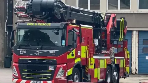 A red and yellow fire engine, facing the camera. Behind it is a grey block of flats with a blue door. There are windows on the second floor. 