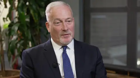 A man, Richard Fuller, is sat wearing a suit, with white shirt and a blue tie