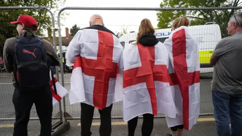 PA Media Five people, three of whom have the flag of St George draped round their shoulders, standing behind a metal fence. In front of them is The Bell Hotel, which is blocked off by police vans.