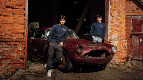 Crossley Motorsport Two young men stand in front of a dilapidated red Aston Martin in a red-brick garage.