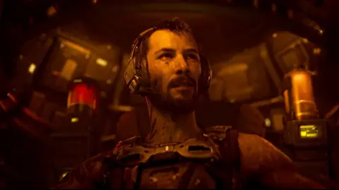 Prime Video A battle-scarred man sits inside the futuristic cockpit of a robot craft, illuminated in a dim orange light emanating from the controls. He wears a communications headset and a metal harness.