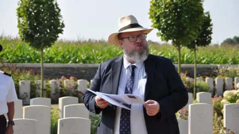 Commonwealth War Graves Commission Peter Francis, a bearded man with glasses and wearing a suite and a sunhat, reading from a paper programme. He's standing in front of rows of white graves and it's a sunny day.
