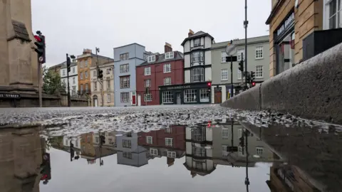 Lucie Johnson A puddle in the foreground reflects the image of the row of traditional colourful buildings in Oxford city centre. The image is taken at ground level with the kerb seen to the right of the shot. The grey skies can be seen above the buildings