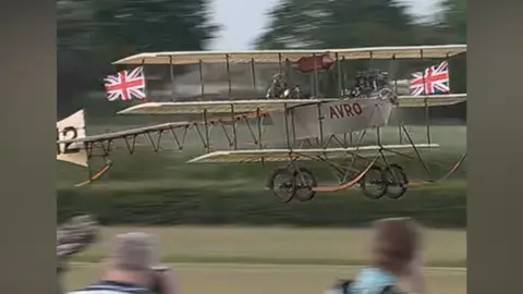 An aircraft taking off as part of the Shuttleworth air show. There are flags on the plane and the word AVRO is on the side of it in red lettering.