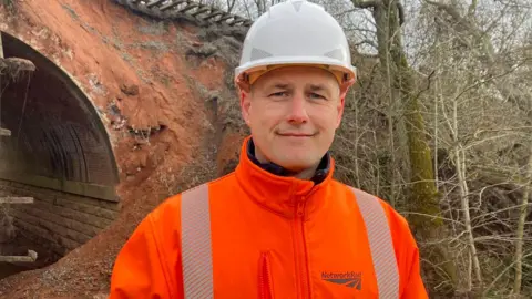 A man wearing an orange hi-vis jacket reading "Network Rail" and a white hard hat, stood in front of a bridge and trees