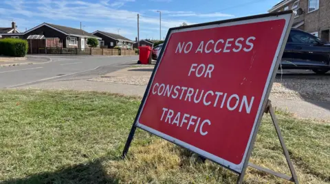 Ben Schofield/BBC A temporary rectangular roadsign that reads "no access for construction traffic" in white text on a red background. It is standing on a grass verge by the side of the road. Two bungalows can be seen in the background.