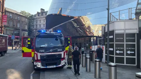 A fire engine stands outside New Street Station, with fire personnel seen standing by the engine. 