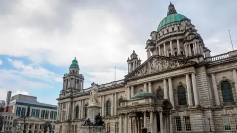 Getty Images A picture of Belfast City Hall. It is a grand building with faded white stone and three towers each of which have a turquoise metal roof.