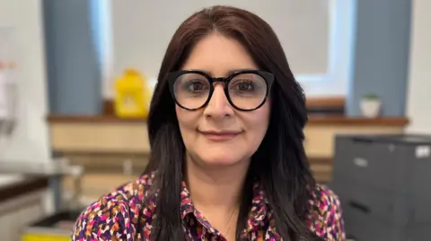 Shabina Qayyum sitting at a desk looking directly at the camera and smiling. She is wearing a pink, purple and gold patterned shirt. She is also wearing a pair of black glasses on her eyes.