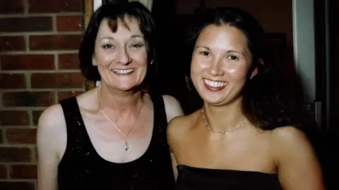 Submitted Two women standing side by side. Both have short dark hair and are wearing black outfits. They are standing outside, in front of a house.