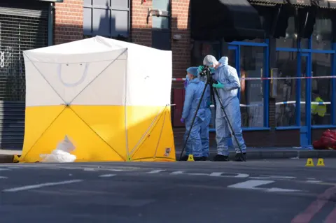 PA Media Two forensic officers dressed in blue hazmat suits take photos next to a white and yellow police tent and cordon