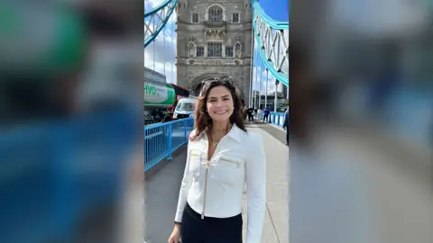 Valentina Gomez has long dark hair, is wearing a white jacket and black trousers, standing on Tower Bridge.