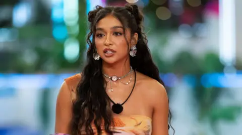 Cierra Ortega on Love Island USA. Cierra has long, wavy dark hair and brown eyes. She wears a patterned one-shouldered orange dress and a microphone around her neck. 
