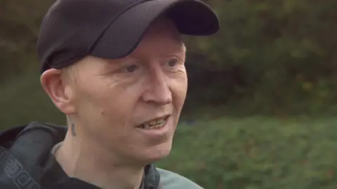 Christopher Crook, wearing a black hat and grey hoodie talking to a reporter outside Forest Bank prison