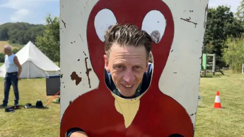 Matt Larcombe. A man is stuck in stocks, namely a board chick, with his head poking through. He has wet brown hair and is smiling. Behind him is a green field. 