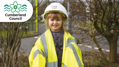 A little girl with blonde hair wearing a yellow hi-vis jacket which is too big for her and a white hard hat. She's smiling into the camera and is standing outside in front of some trees and a road.