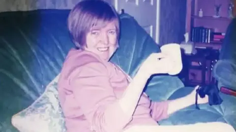 Family photo Judith Hindley is sitting on a blue sofa and is holding a mug. She is wearing a pink top and is looking at the camera side on and smiling