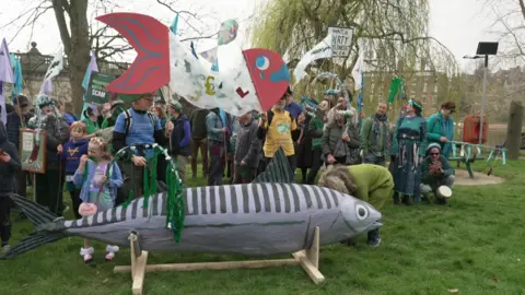 BBC A large grey model fish can be seen in front of about 40 to 60 people, some waving banners and placards, by the river. They are standing on grass.
