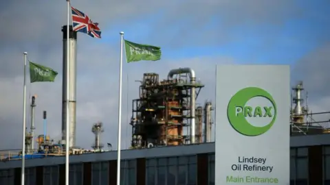 Getty Images A metal structure in the background with a board on the left reading: 'PRAX. Lindsey Oil Refinery. Main Entrance'. There is a union jack flag on the left between two green flags with PRAX written.