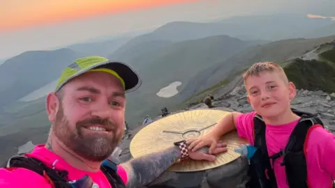 Daniel Thomas and Albie-Junior standing at the peak of Yr Wyddfa, also known as Snowdon. An orange and pink sunset can be seen in the horizon. They are both smiling and looking at the camera.