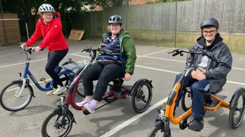 James Grant/BBC Three students sit on three "trikes". All three are wearing helmets and are smiling at the camera. 