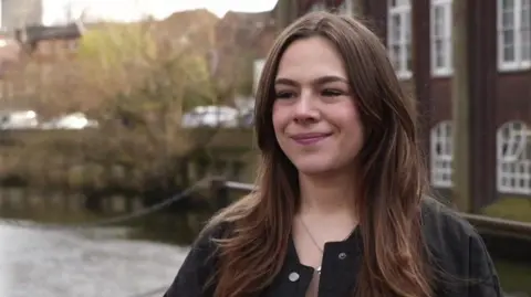 Shaun Whitmore/BBC Josephine Willis, a student who is standing outside with the River Wensum in the backdrop. She is looking to the left of the camera and is smiling. She is wearing a black jacket and has brunette hair.