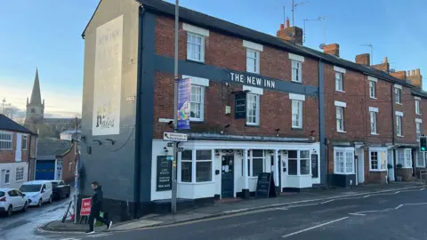 Tony Fisher/BBC A street scene in Buckingham. There is a church in the background and the New Inn in the foreground. It is at the end of a row of traditional buildings  with red bricks and white sash windows.